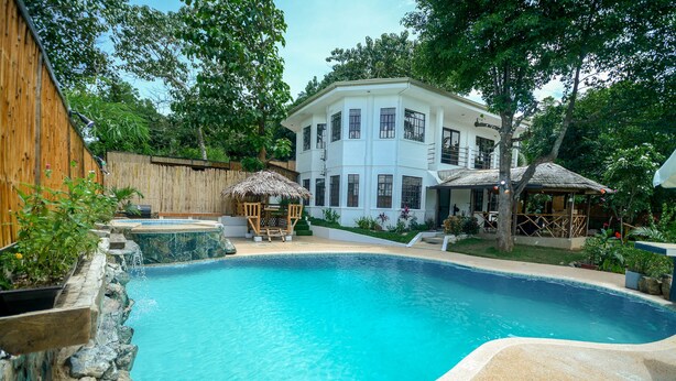 A swimming pool with a stone wall, a wooden fence, and a white building with multiple windows. - Hilltop Pool and Villa (Puerto Princesa)