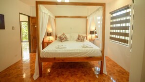 A four-poster bed with white linens and decorative pillows, a wooden floor, and a window with blinds. - Hilltop Pool and Villa (Puerto Princesa)