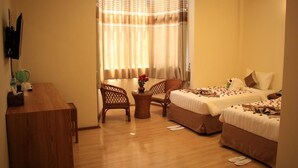 Una habitación de hotel con dos camas, un escritorio de madera, una mesita con un jarrón de flores y un televisor montado en la pared.