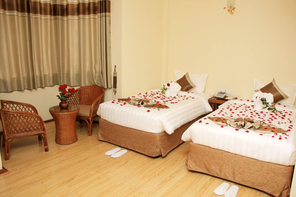 A hotel room with two beds, a wicker chair, a small round table, and a pair of slippers on the floor.