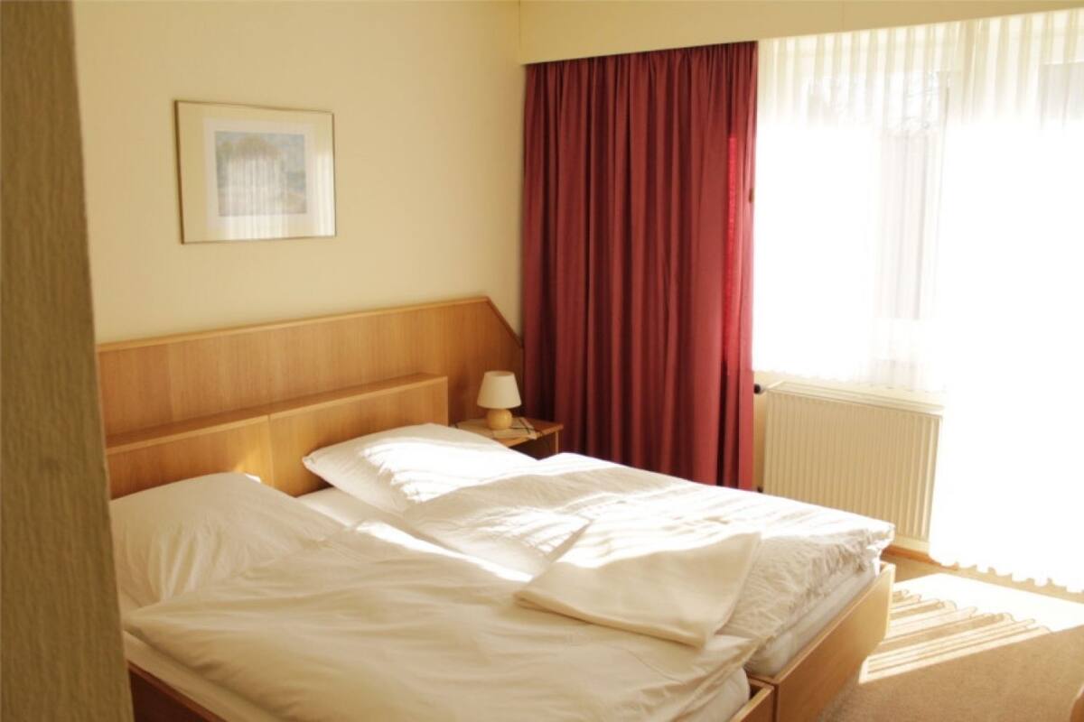 Una habitación de hotel con cabecera de madera, una cama individual con ropa blanca, una mesita de noche con lámpara, un cuadro enmarcado en la pared y una ventana con cortinas rojas.