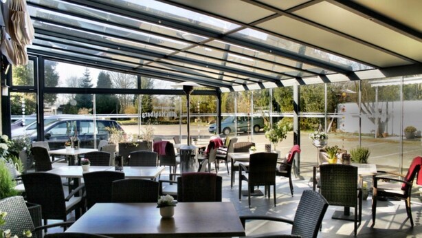 Un café al aire libre con mesas y sillas, una estructura de vidrio y vista a la calle.