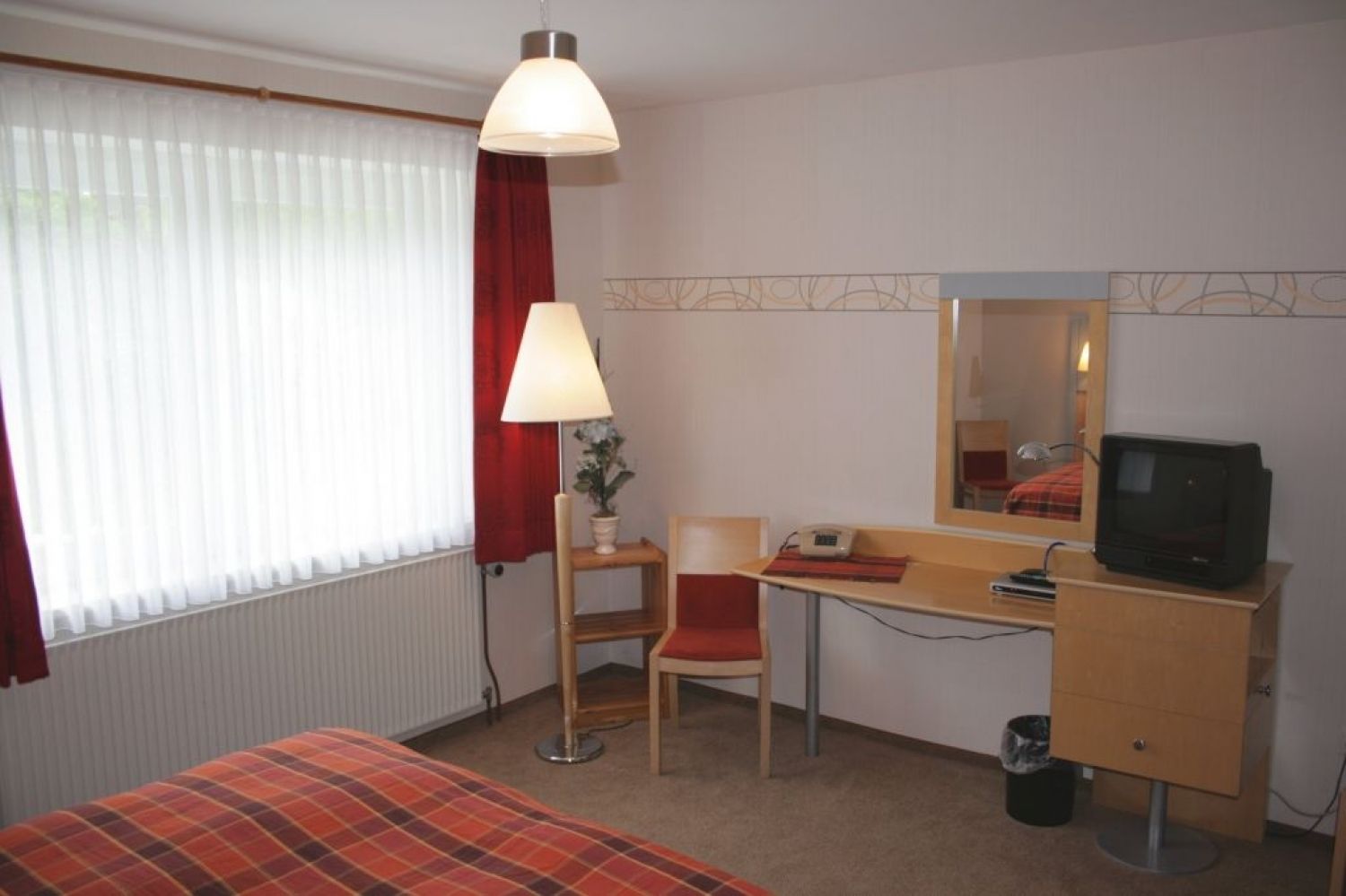 A hotel room with a bed, a desk with a television, a chair, a lamp, and a window with curtains.