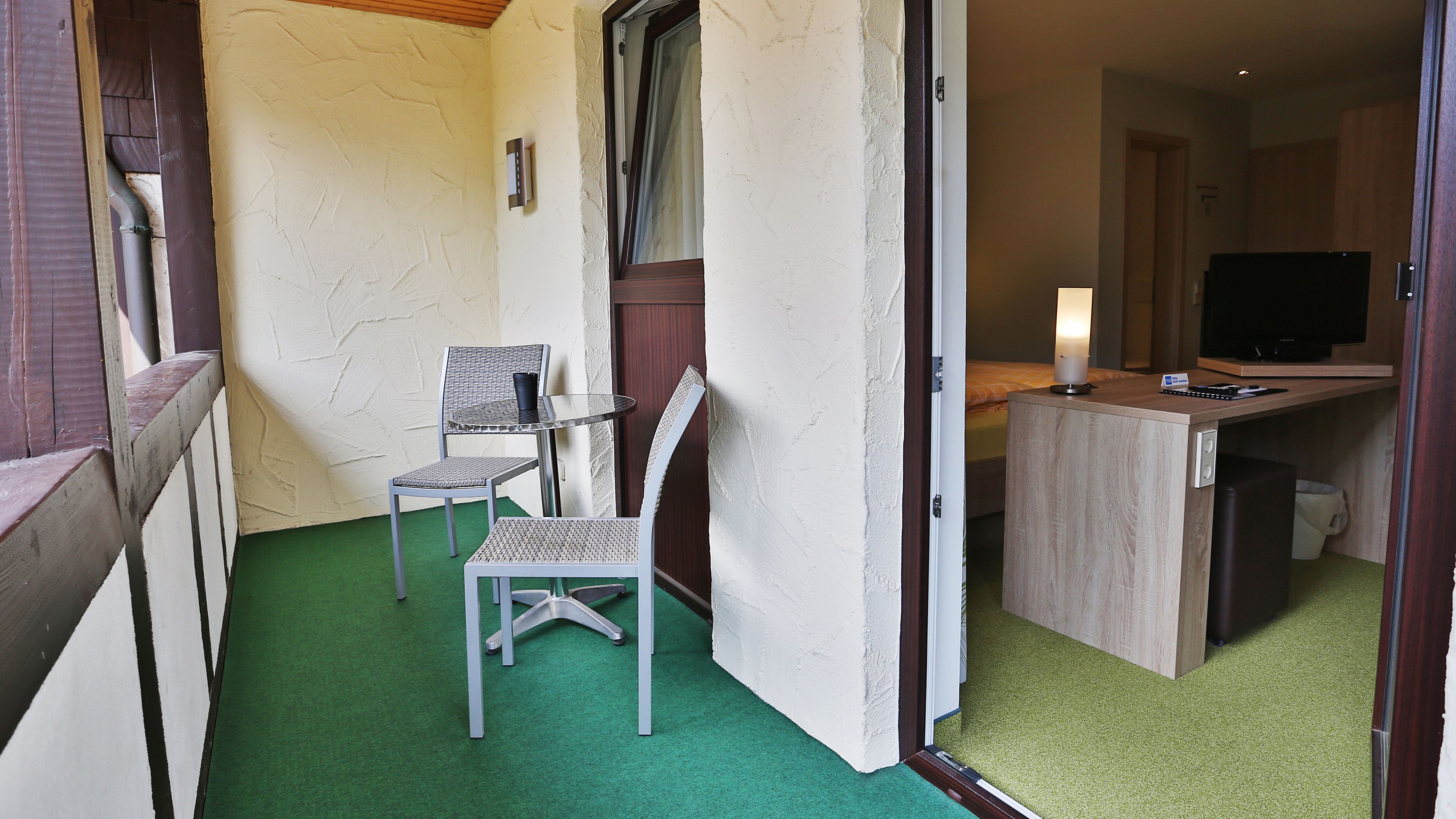 Ein Hotelzimmer mit grünem Teppich, zwei Korbstühlen, einem kleinen Tisch, einem Bett, einem Fernseher und einem Schreibtisch mit Lampe.