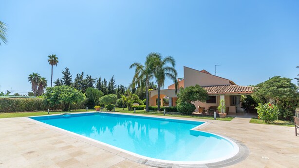 A rectangular swimming pool with a tiled deck, surrounded by palm trees and a house with a tiled roof. - Villa Arenella con Piscina by Wonderful Italy (Syracuse)