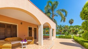 A covered outdoor seating area with wicker furniture and a small table, surrounded by palm trees and a clear blue sky. - Villa Arenella con Piscina by Wonderful Italy (Syracuse)