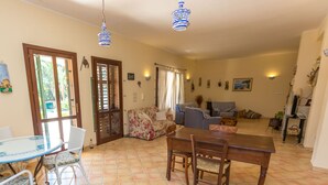 A spacious living area with a dining table, chairs, a sofa, and a coffee table. There are two doors leading to a balcony, and the room has tiled flooring and ceiling lights. - Villa Arenella con Piscina by Wonderful Italy (Syracuse)