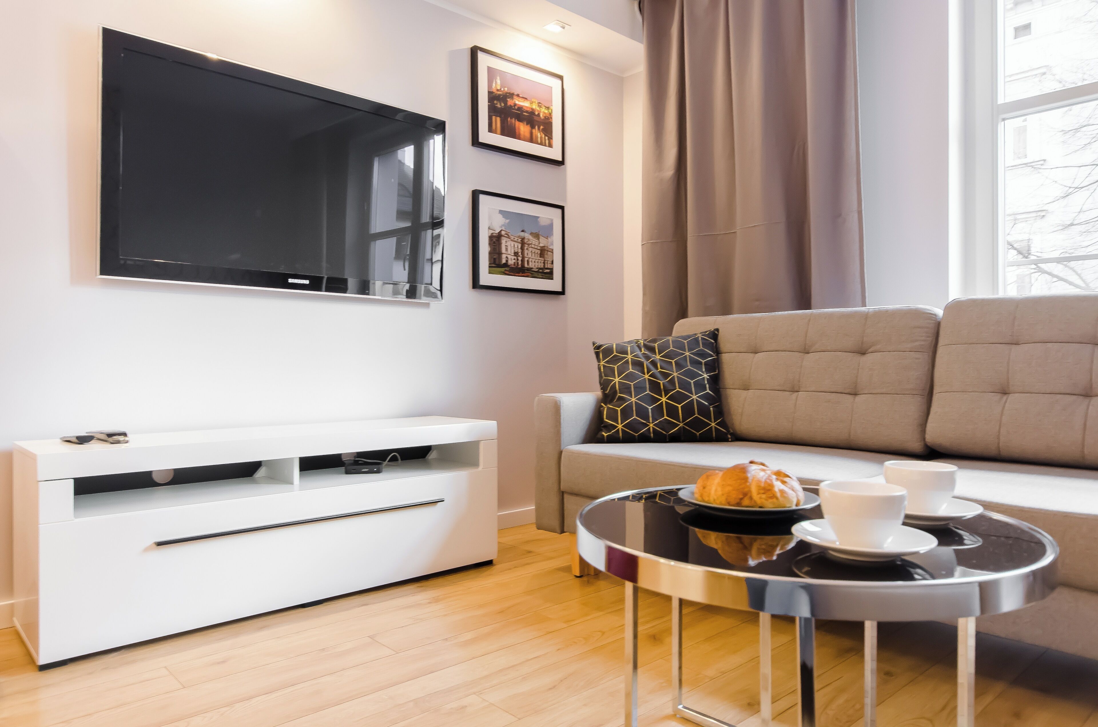 A modern living room with a flat-screen TV, a sofa, a coffee table with a croissant, and two cups.