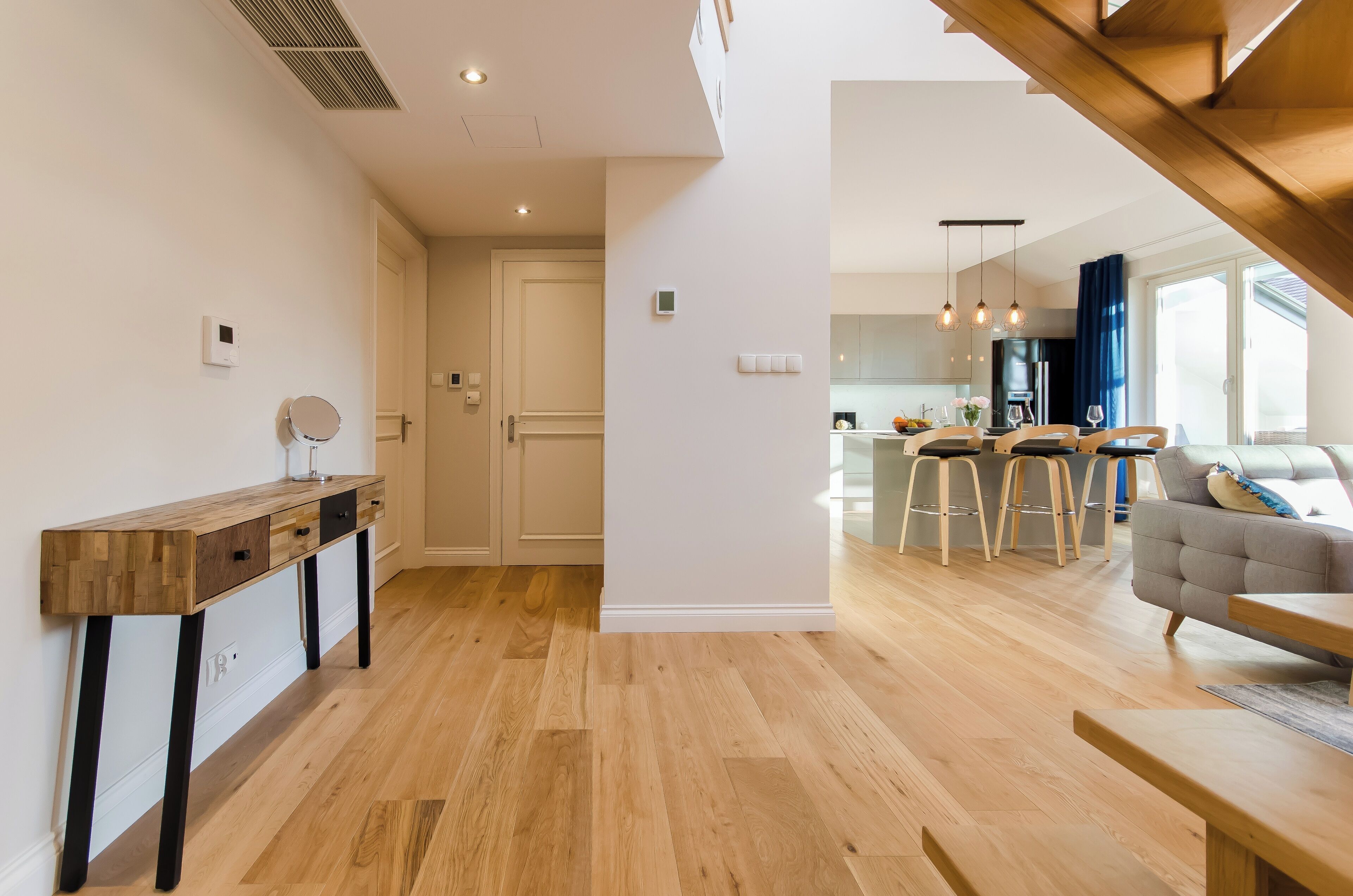 A modern interior with a wooden console table, a kitchen area with bar stools, and a grey sofa.