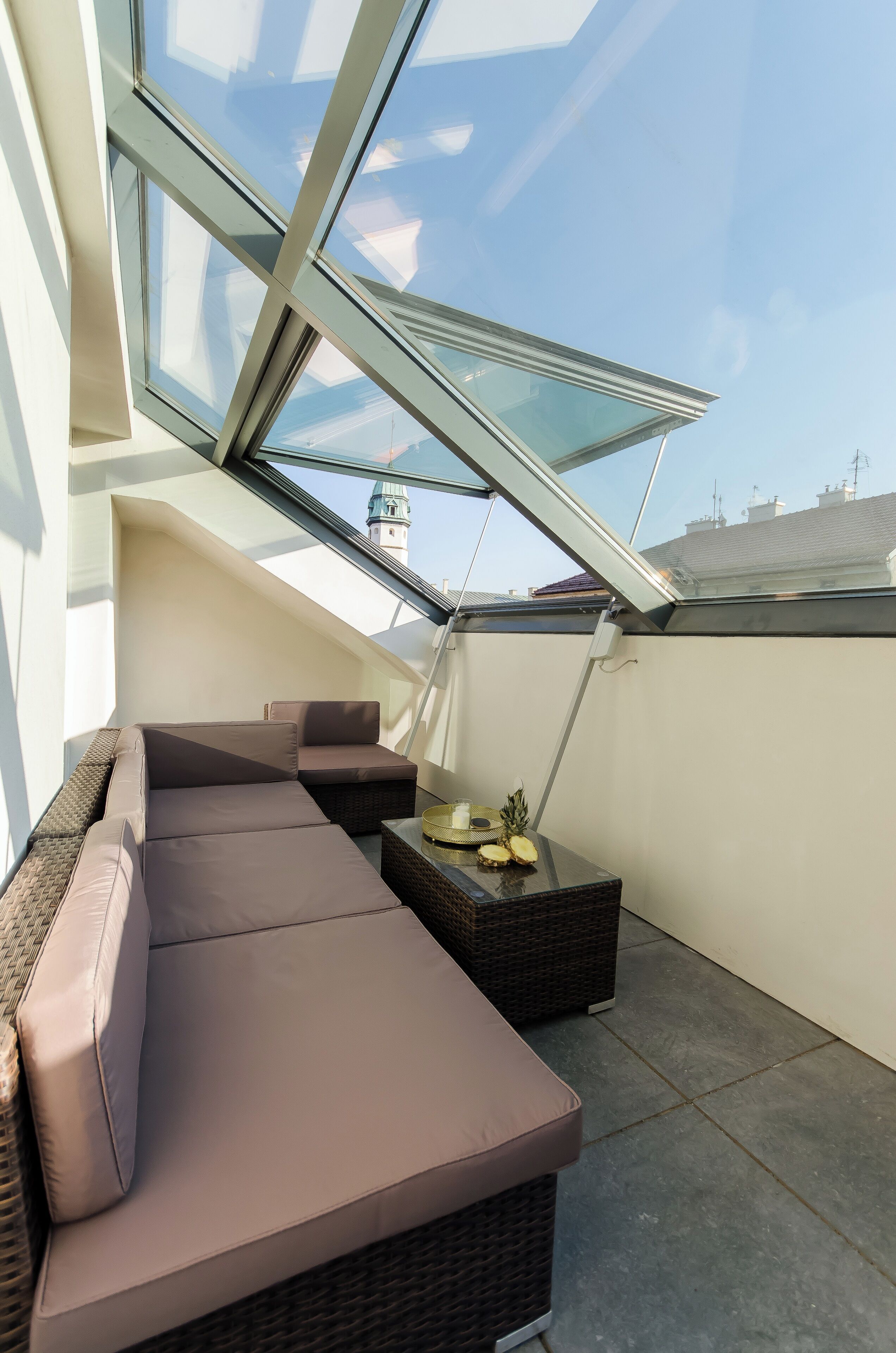 Una terraza en la azotea con techo de vidrio, un sofá de mimbre marrón y una pequeña mesa con una piña.