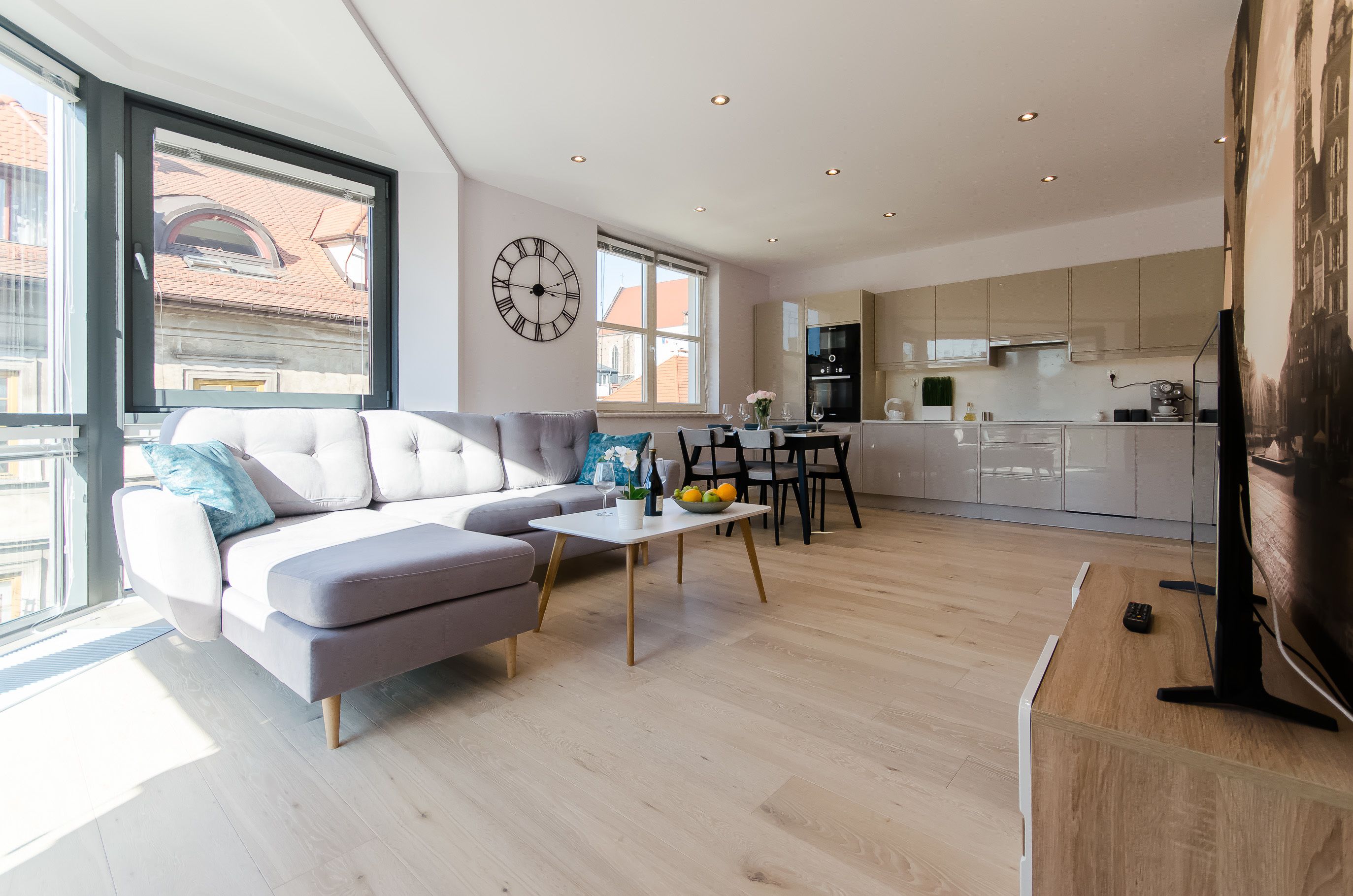 En moderne stue med en grå sofa, et spisebord med stole, et køkkenområde og et stort vindue med et ur på væggen.