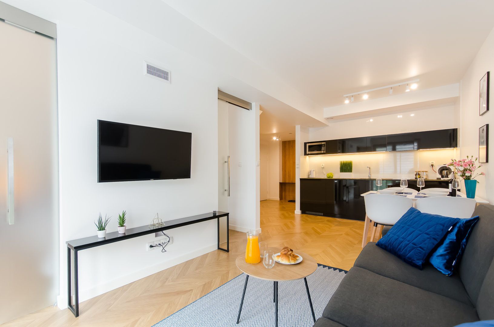 Un salon moderne avec un téléviseur à écran plat, une console agrémentée de plantes, un canapé gris avec un coussin bleu et un coin repas avec une cuisine en arrière-plan.