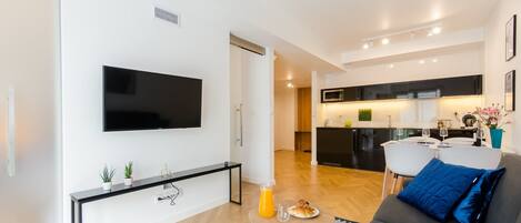 Un salon moderne avec un téléviseur à écran plat, une console agrémentée de plantes, un canapé gris avec un coussin bleu et un coin repas avec une cuisine en arrière-plan.