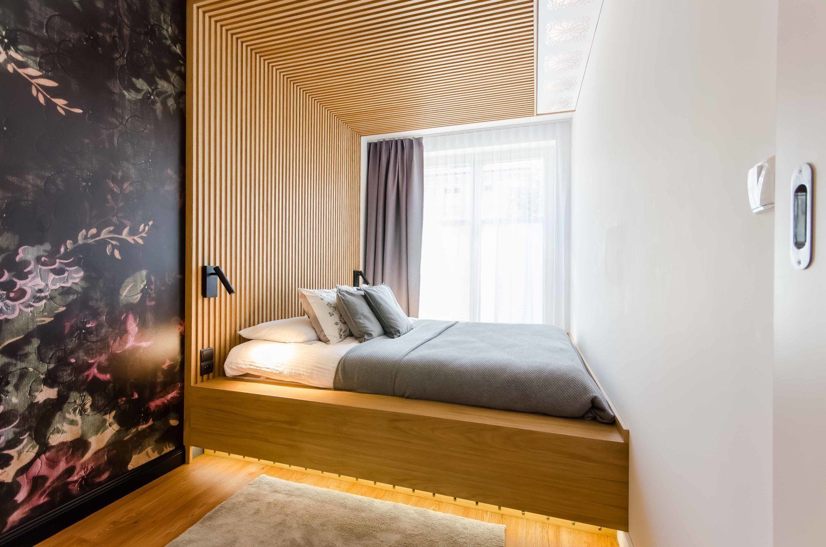 Une chambre moderne avec un plafond en pente, un lit intégré, une grande fenêtre avec des rideaux et un luminaire fixé au mur.