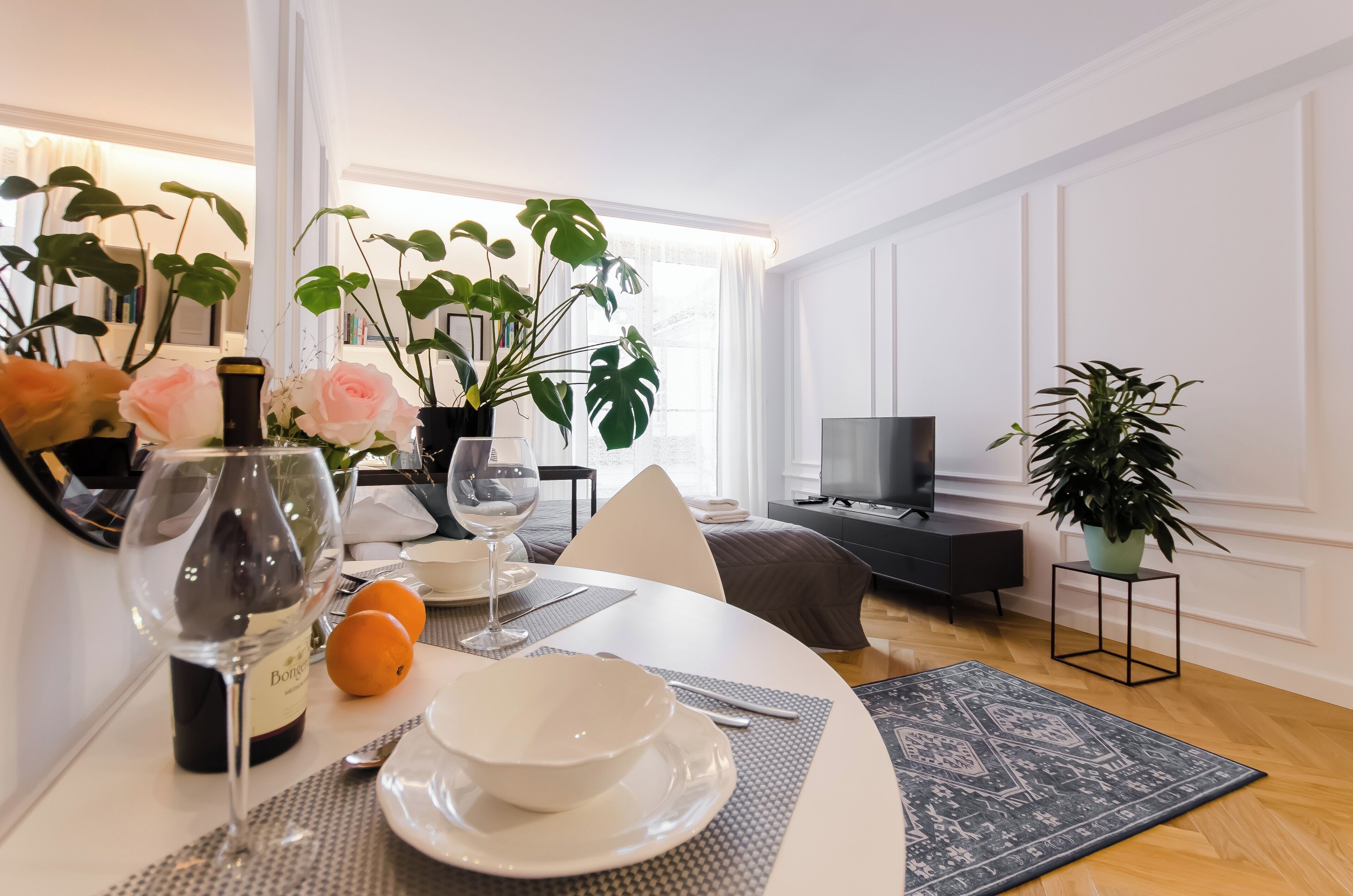 A modern, well-lit room with a dining table set for two, featuring a wine bottle, glasses, and a bowl. There are potted plants and a TV in the background.
