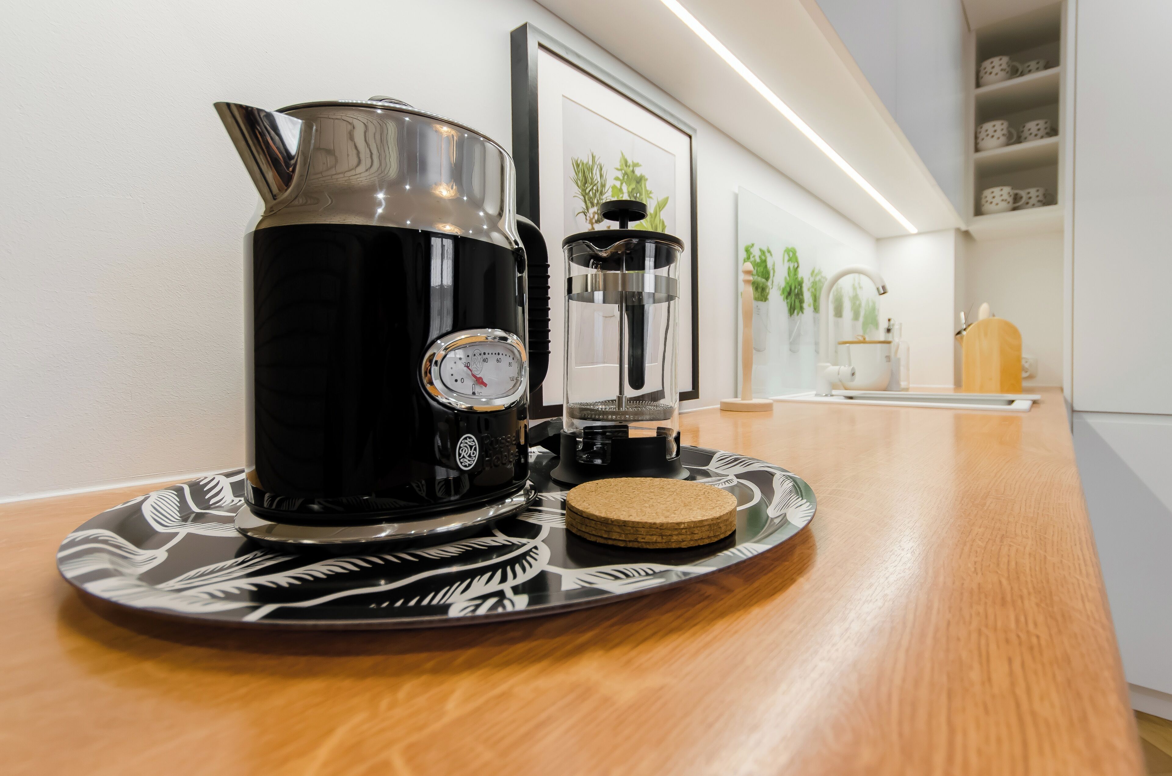 A coffee maker and a grinder on a wooden table with a coaster, framed artwork, and shelves with cups in the background.