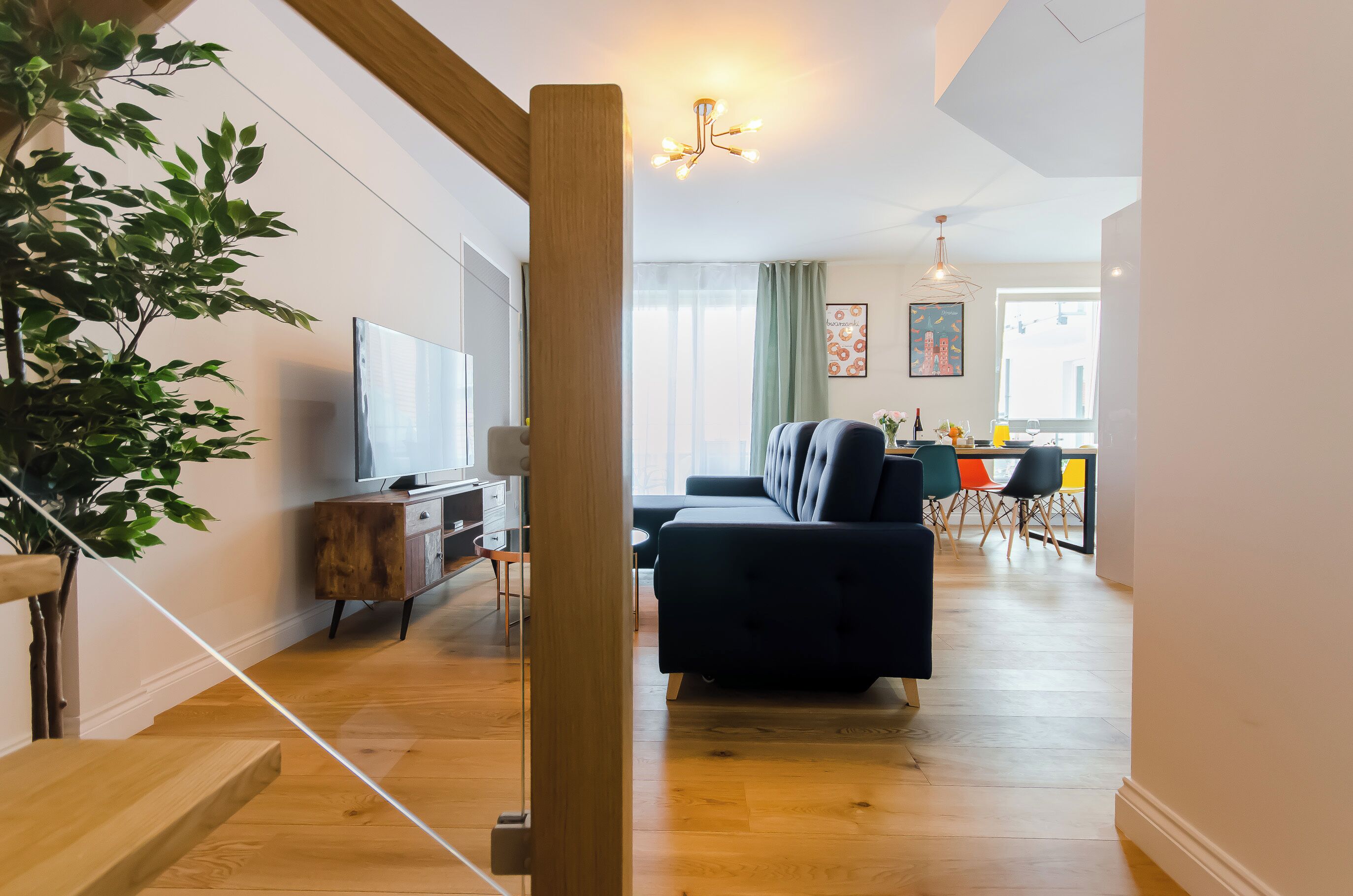 Una sala de estar moderna con un sofá negro, piso de madera, televisor y comedor con sillas coloridas.