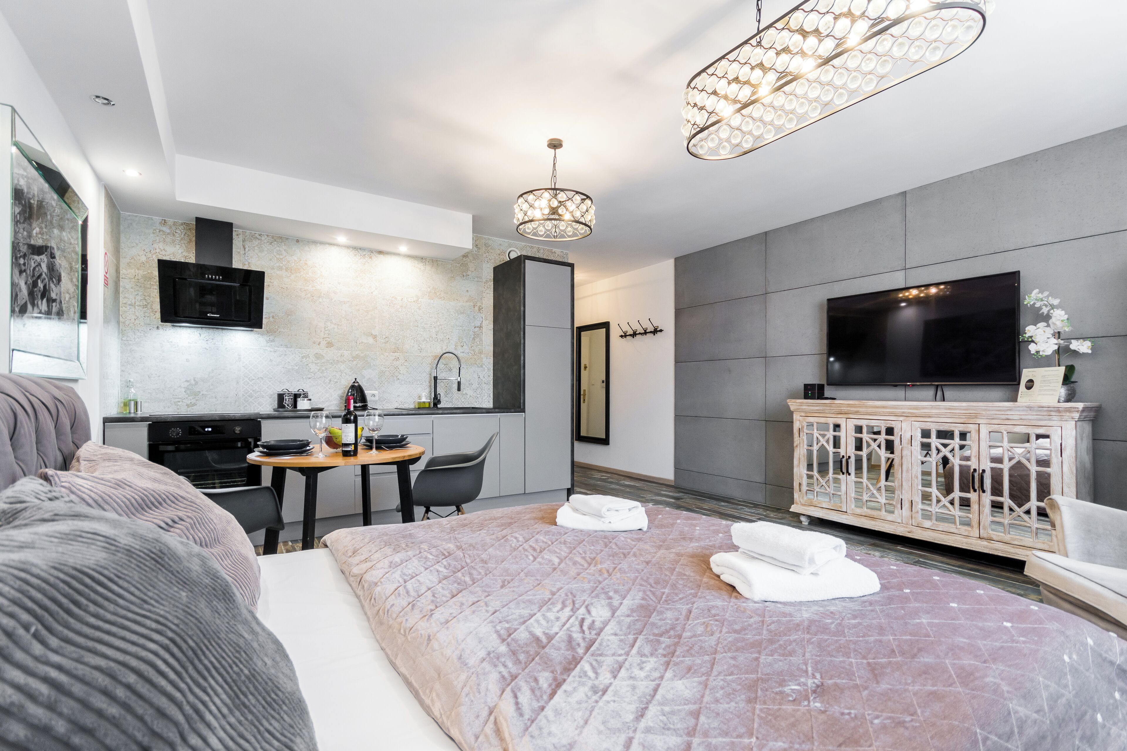 A modern hotel room with a large bed, a dining area, a kitchen, and a TV.