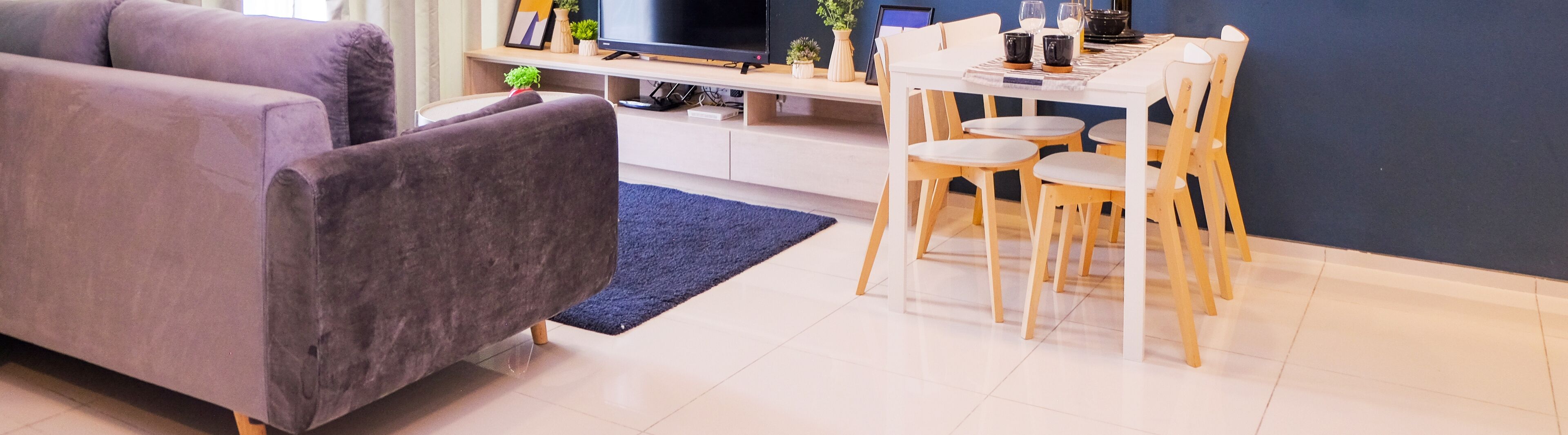 A modern living room with a grey sofa, a wooden dining table with four chairs, a flat-screen TV, and a small plant on the table.