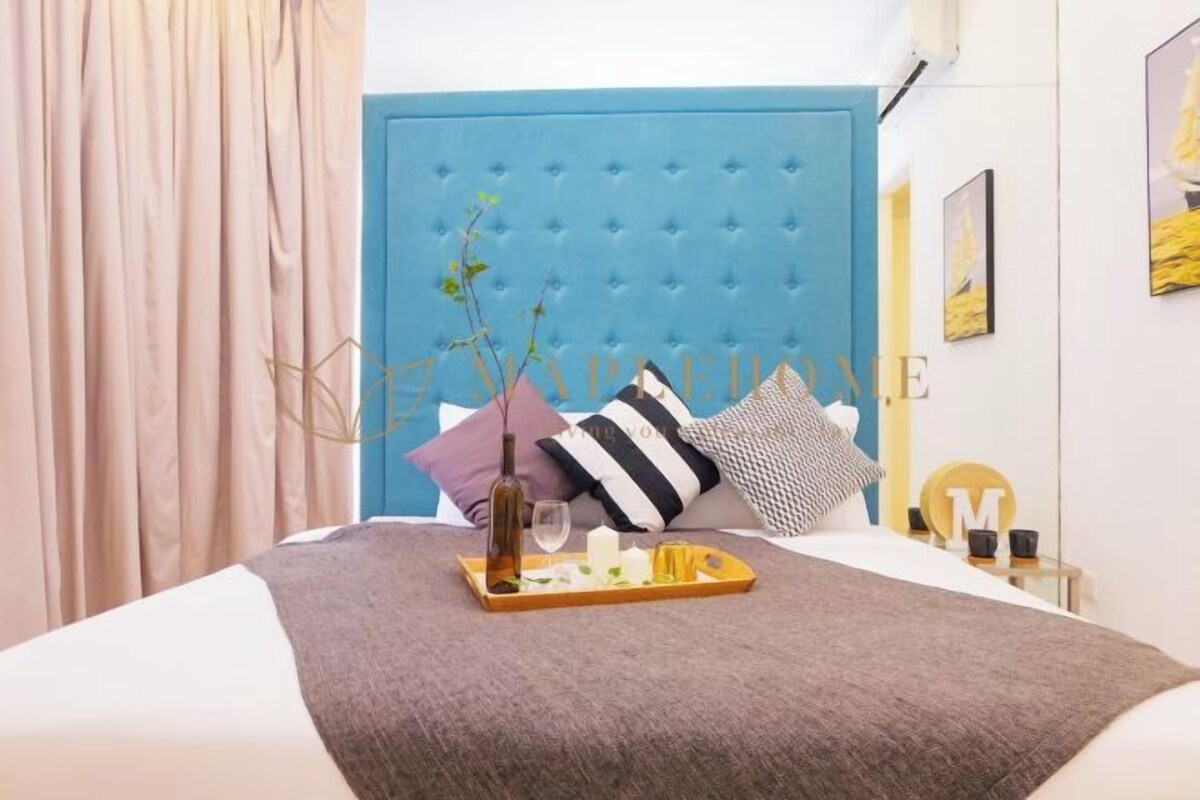 A neatly made bed with a blue headboard, a tray with a bottle and glasses, and a vase with flowers.