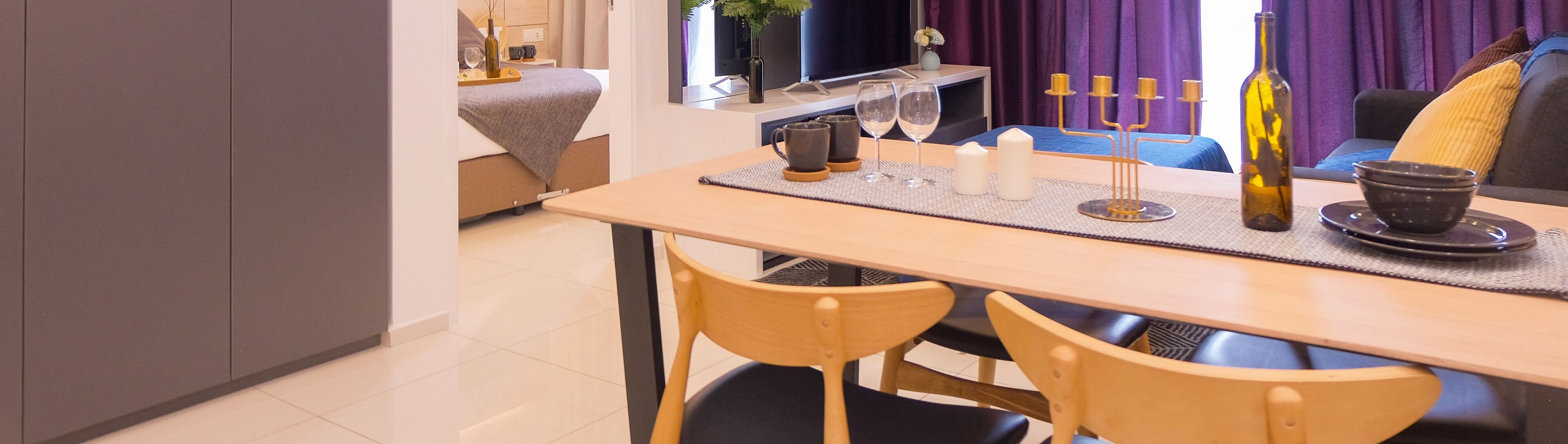 A dining area with a table set for two, a television in the background, and a sofa with cushions.