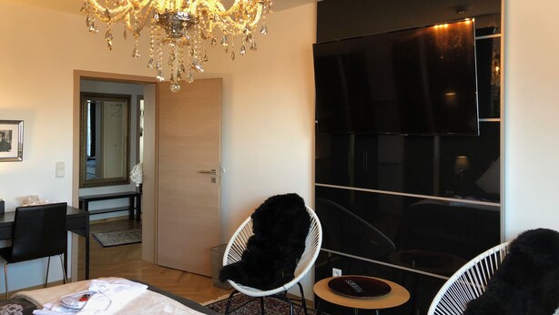 A modern hotel room with a chandelier, a large flat-screen TV, two white chairs, a bed with a floral bedspread, and a wooden floor. - City Apartments Will (Emmerich)