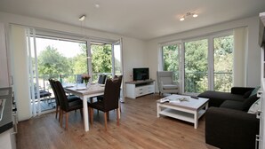 Een ruime woonkamer met een eettafel, stoelen, een bank en een televisie. Grote ramen bieden natuurlijk licht en uitzicht op groen buiten.