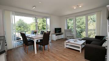 A spacious living area with a dining table, chairs, a sofa, and a TV. Large windows provide natural light and views of greenery outside.