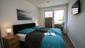 Een slaapkamer met een bed, nachtkastje, lamp, schilderij en televisie aan de muur.