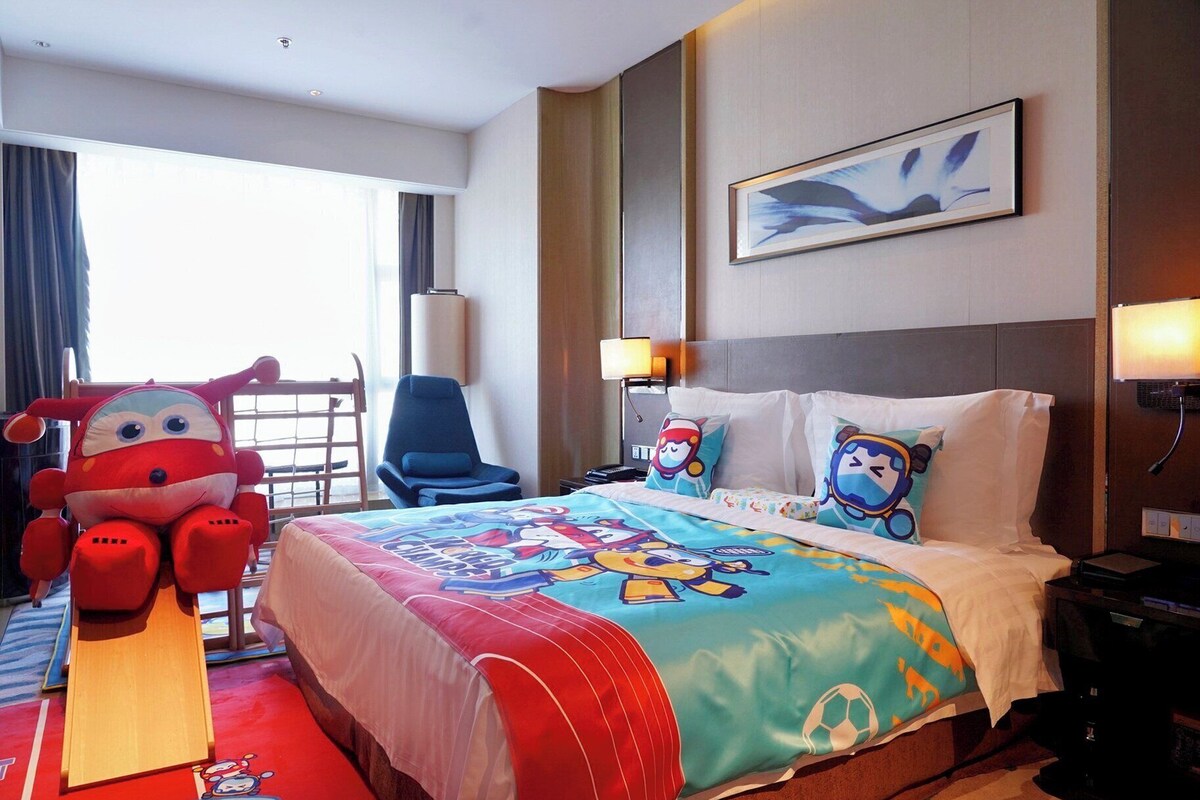 A hotel room with a bed, a chair, a large plush toy, and a painting on the wall.