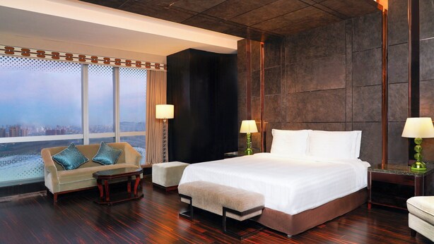 A modern hotel room with a large bed, a sofa, a bench, and a view of the city through large windows.