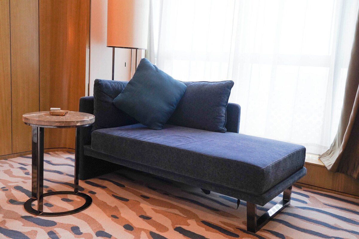 A single sofa with two pillows, a small round table, and a patterned carpet in a room with wooden paneling and a window with curtains.