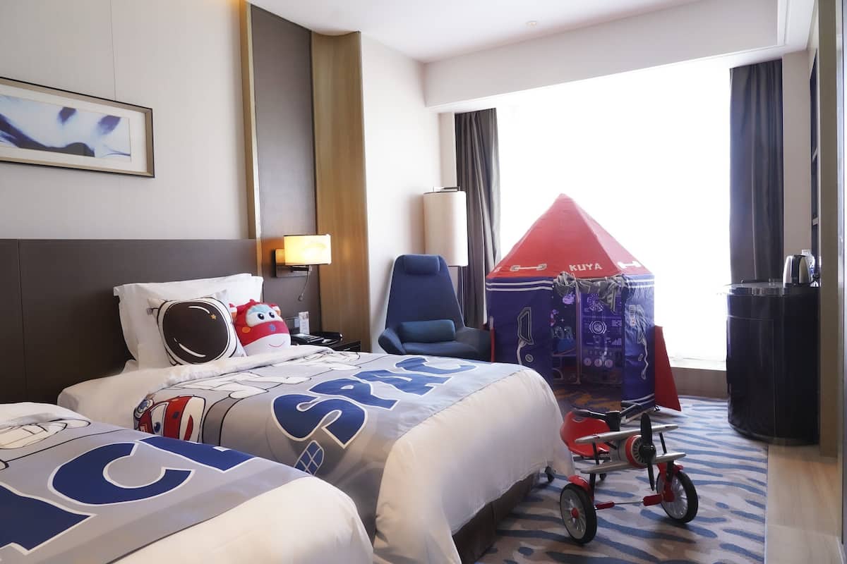 Ein Hotelzimmer mit zwei Betten, einem roten Buggy mit Baldachin, einem blauen Sessel und einer Lampe.