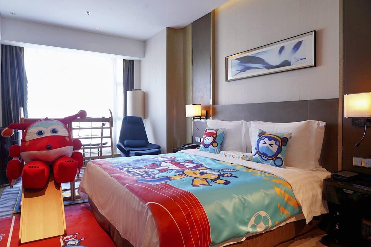 Ein Hotelzimmer mit Bett, Stuhl, einer großen Stoffpuppe und einem Bild an der Wand.