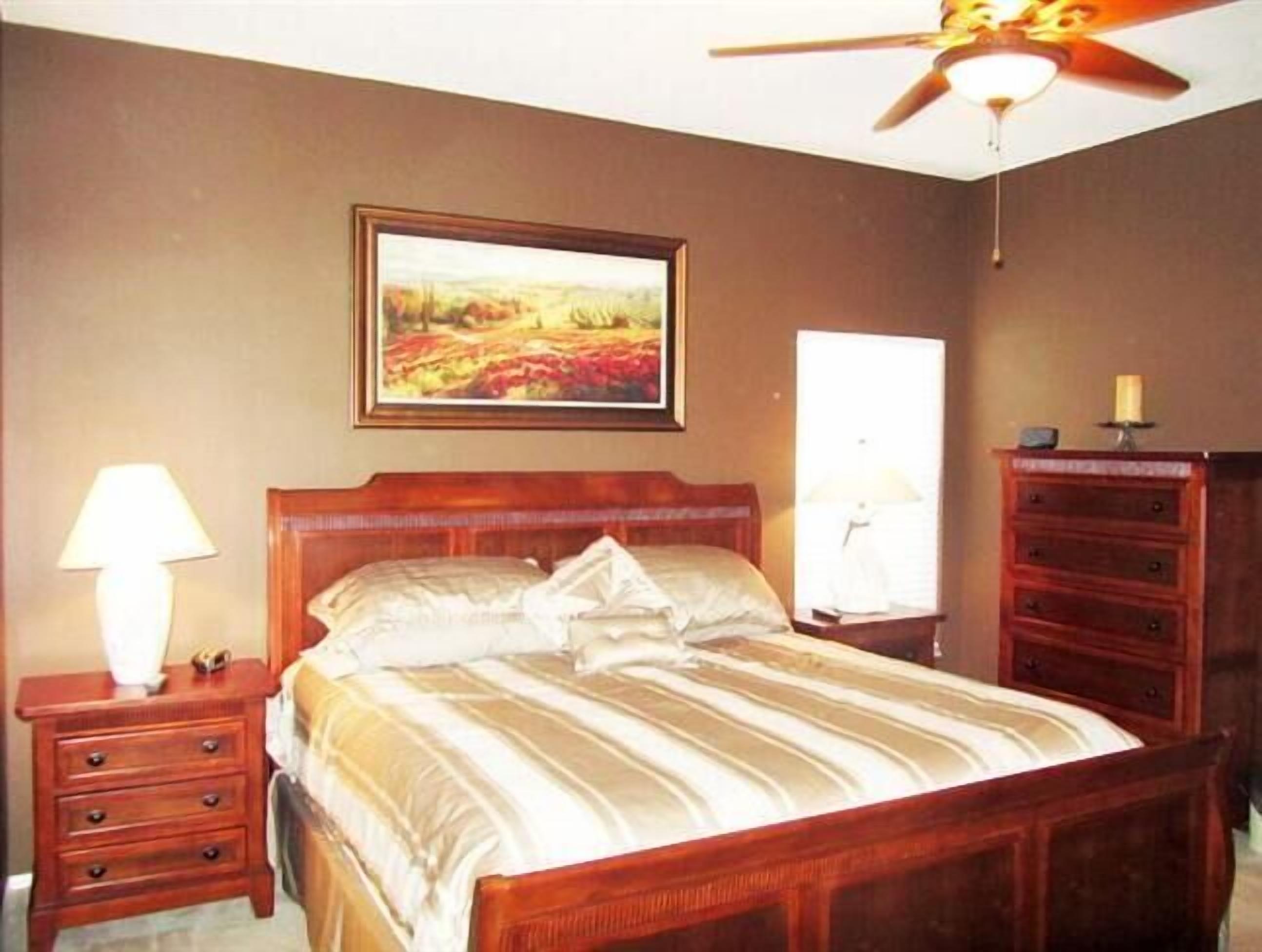 Une chambre à coucher comprenant un lit, une table de chevet, une commode, un ventilateur de plafond et un tableau accroché au mur.