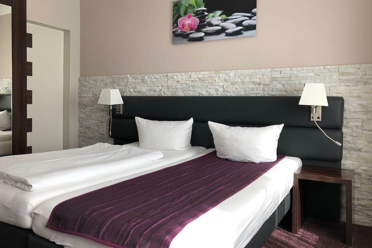 A hotel room with a bed, two bedside lamps, a stone wall, and a painting of flowers and stones.
