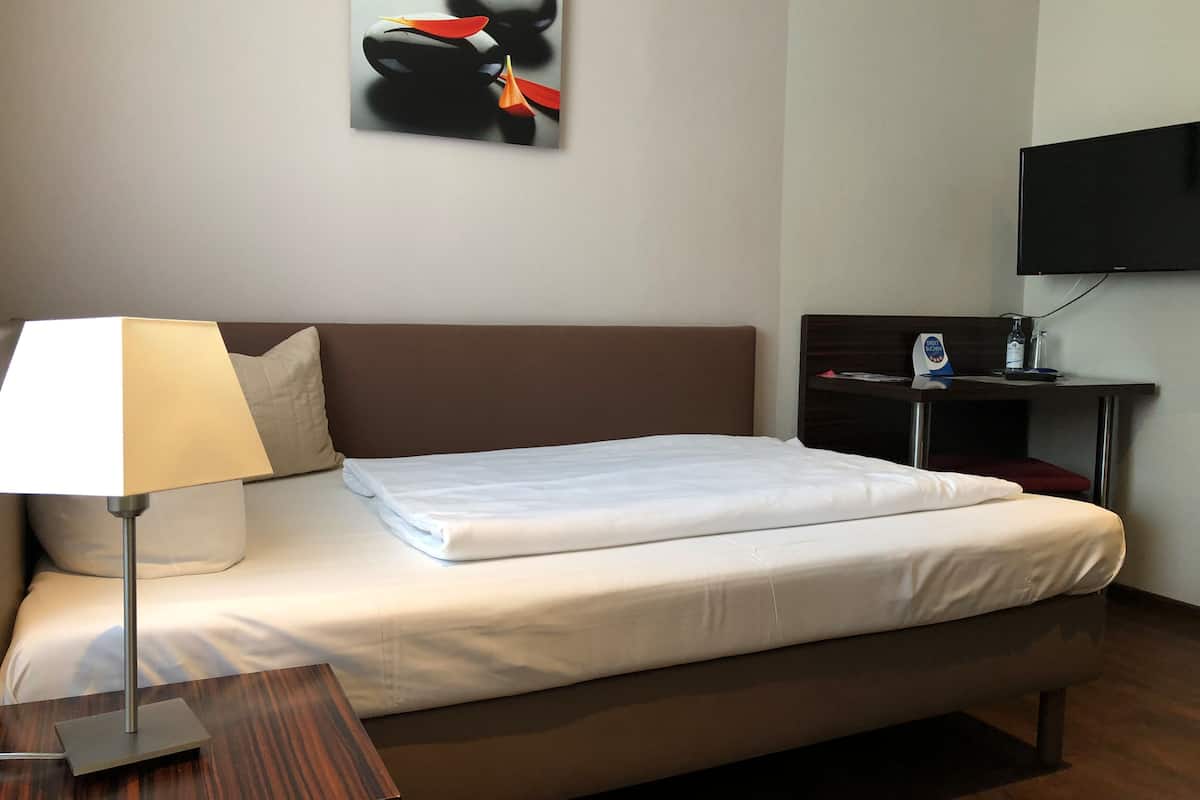 A hotel room with a bed, a bedside table with a lamp, a TV on a stand, and a wall-mounted artwork.
