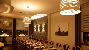 Restaurante com mesas compridas preparadas para refeições, luminárias pendentes e parede de pedra com pintura de moinho.