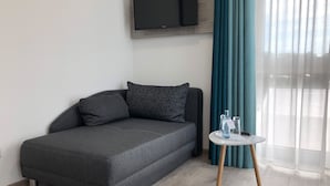 Quarto moderno com sofá cinzento, uma mesa redonda pequena com um tabuleiro, televisão fixada na parede e janela com cortinas azuis.