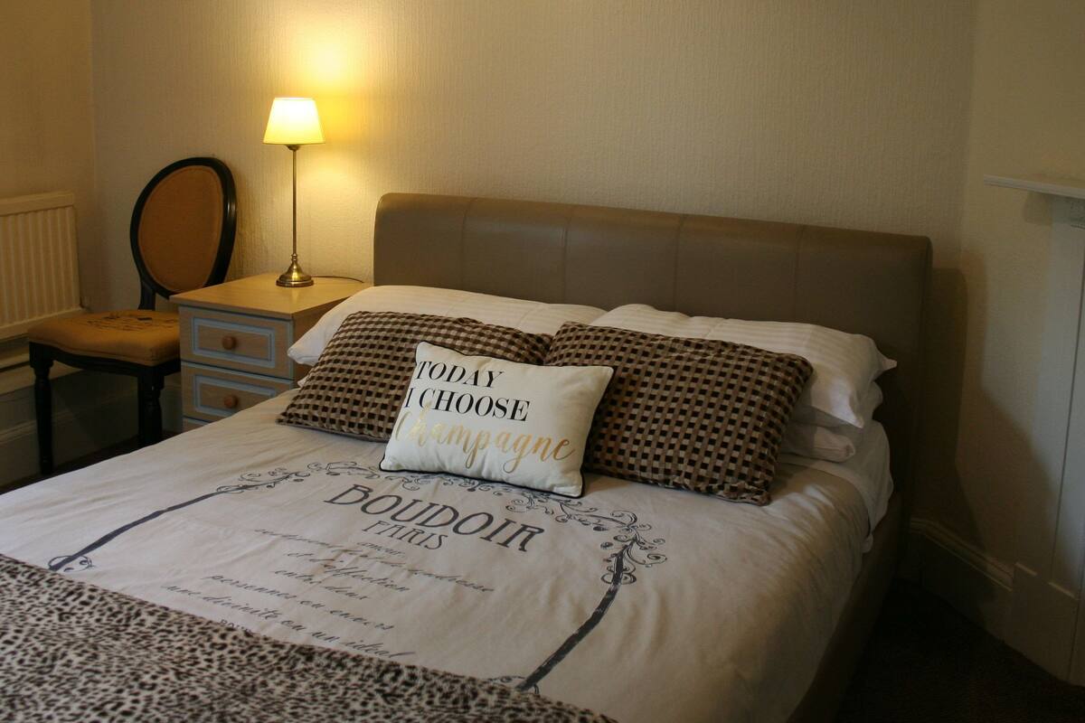 A neatly made bed with a pillow stating "TODAY I CHOOSE Champagne," a lamp, a chair, and a dresser.