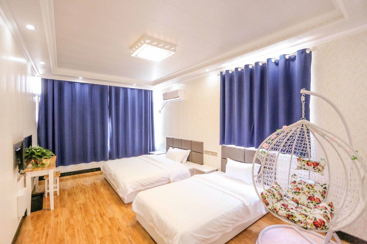 Habitación de hotel con dos camas individuales, un escritorio con televisor y una planta, un sillón colgante y una ventana con cortinas.