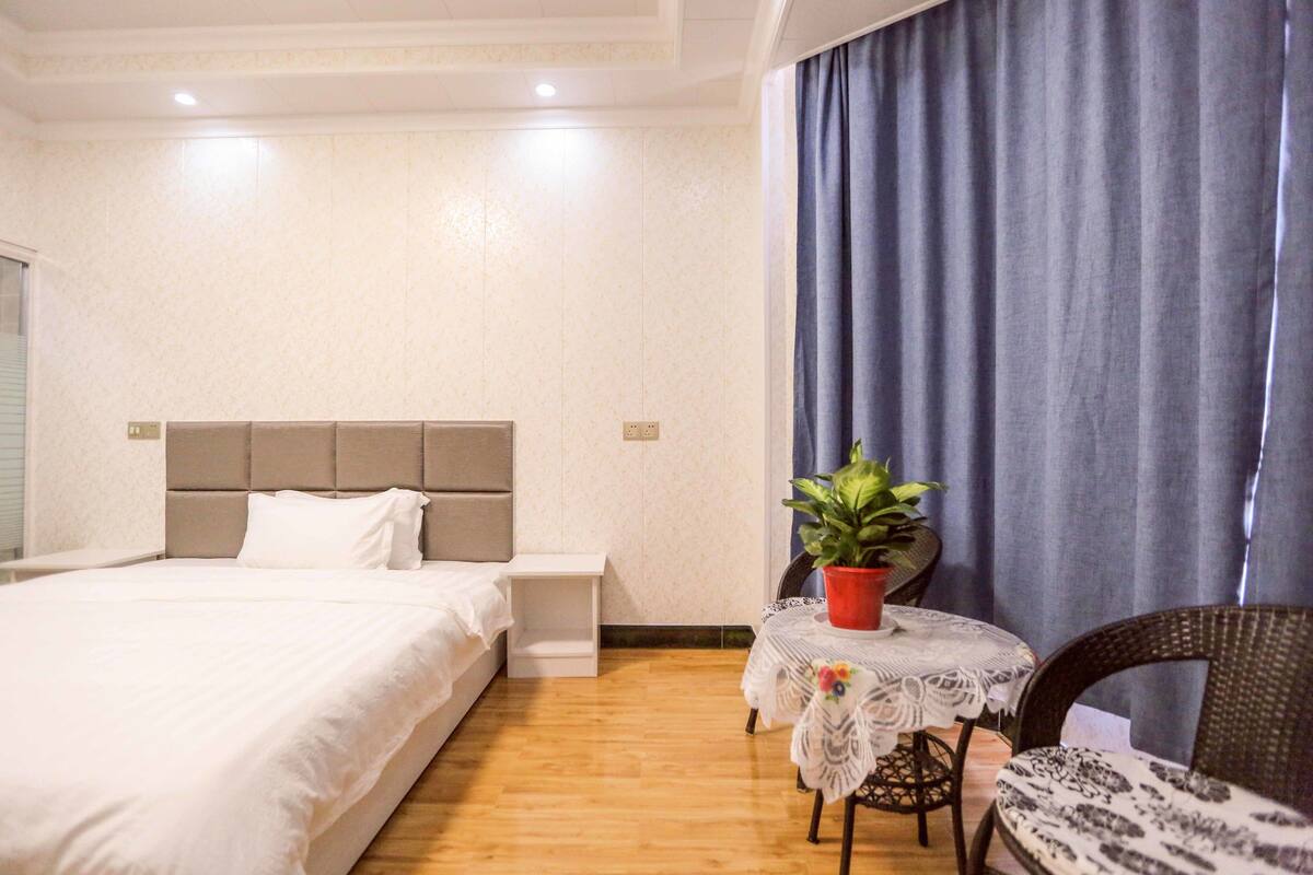 Habitación de hotel con cama, una mesita con una planta, una silla y una ventana con cortinas.