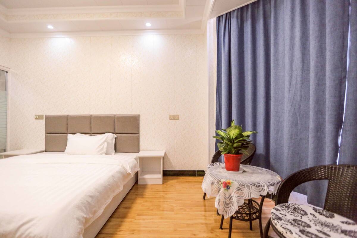 Un dormitorio con cama, mesita de noche, planta en maceta y una pequeña mesa redonda con una silla.