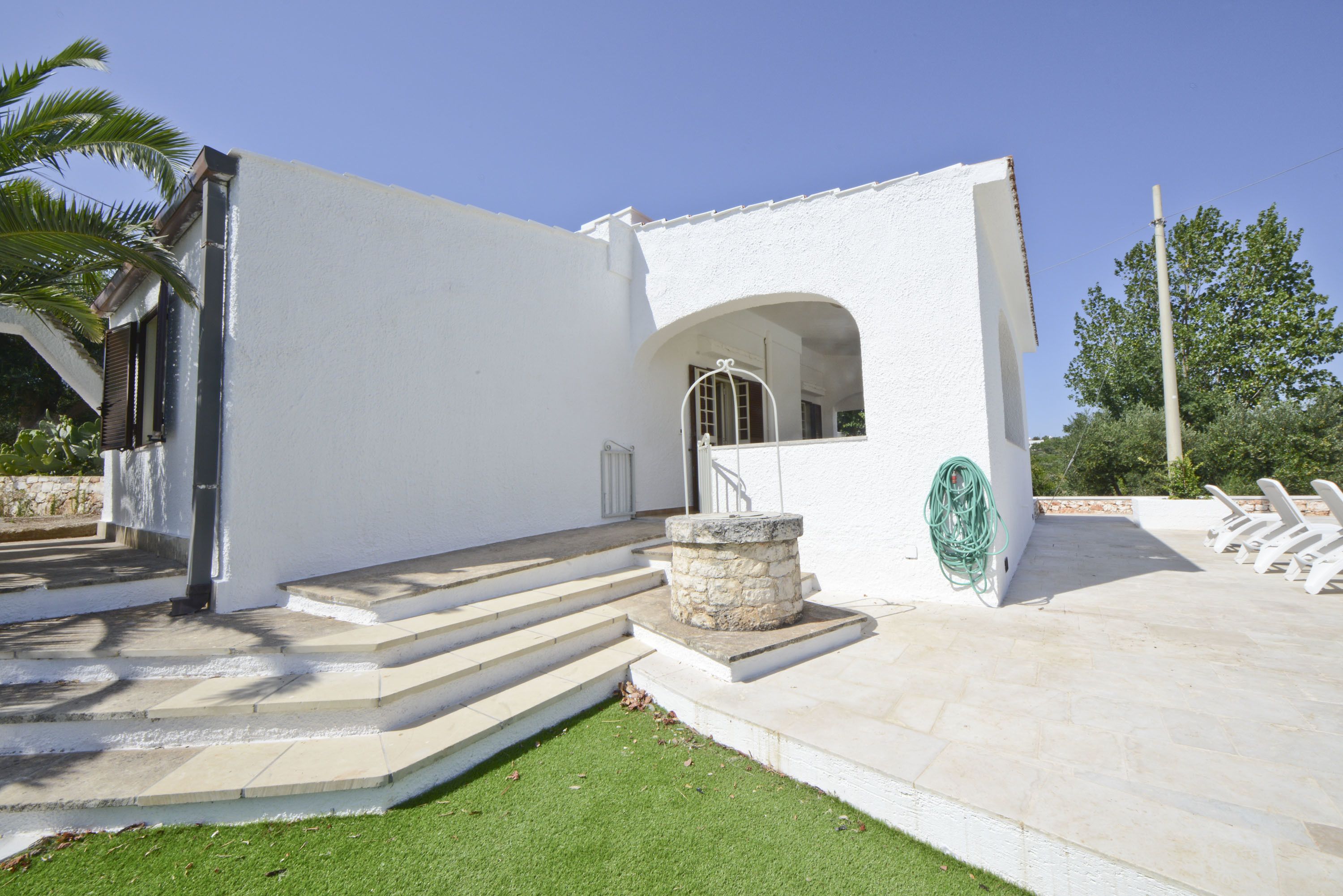 A white building with a stone staircase leading to a door, a stone planter, and a paved patio area with white chairs.