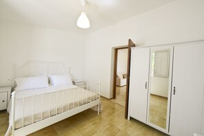 A bedroom with a white bed, a nightstand, a wardrobe, and a door leading to another room. - Villa d'Itria with Trullo by Wonderful Italy (Ostuni)