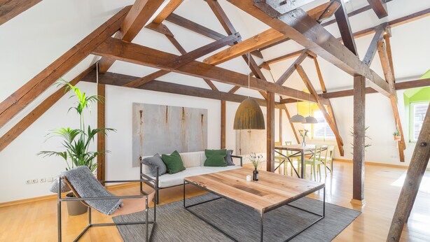 A spacious loft with exposed wooden beams, a wooden dining table, a sofa with green cushions, and a hanging plant.