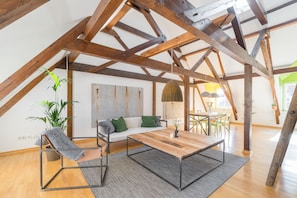 Un loft espacioso con vigas de madera a la vista, una mesa de comedor de madera, un sofá con cojines verdes y una planta colgante.