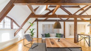 A spacious loft with exposed wooden beams, a wooden dining table, a sofa with cushions, a chair, and a hanging pendant light.