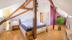A bedroom with a wooden ceiling structure, a bed with white bedding, a bedside table, a potted plant, and a wooden bench.