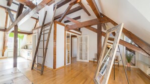 An attic room with exposed wooden beams, a wooden ladder, and a large window.